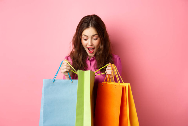 Izgalmas lány kinyit bevásárló táskák és ziháló csodálkozva, ellenőrzi ajándékokat neki boldog arccal, áll szemben rózsaszín háttér - Fotó, kép