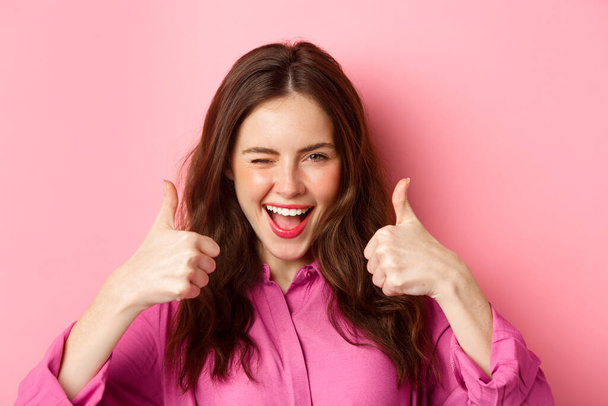 Cheeky glamour girl winking at you and shows thumbs up, praise great work, good choice, standing over pink background - Photo, Image