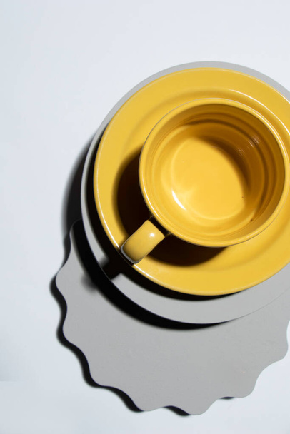 Κίτρινο φλιτζάνι καφέ δίπλα σε γκρι γεωμετρικά σχήματα και σκληρό φως - Φωτογραφία, εικόνα