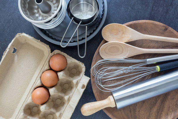 台所用品と成分の概念は、暗い背景に泡立て器、木製のスプーンと新鮮な卵とカップを測定します。 - 写真・画像
