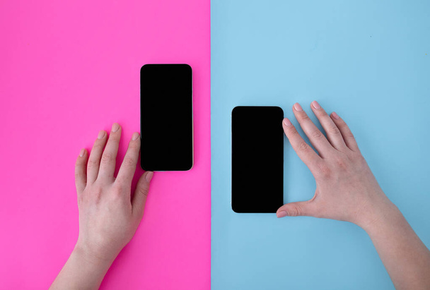 Manos femeninas tocando dos teléfonos inteligentes con pantallas en blanco sobre fondos azules y rosados, vista superior, posición plana - Foto, imagen