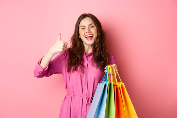 Молода щаслива жінка-покупець, яка показує великі пальці вгору, задоволена хорошими знижками, купує персонал на продаж, тримає сумки для покупок і посміхається задоволений, рожевий фон
 - Фото, зображення