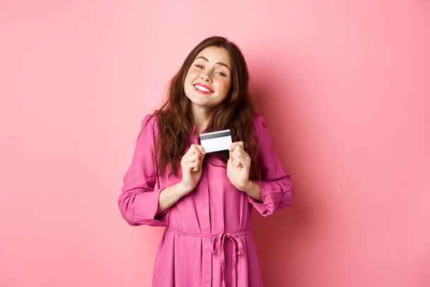 Young brunette woman hugging her credit card with delight, smiling pleased, standing against pink background - Photo, Image