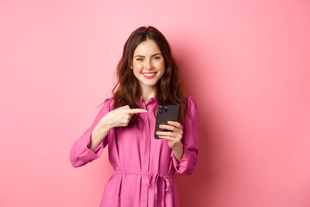 Hübsches kaukasisches Mädchen wirbt für Online-Angebot, zeigt mit dem Finger auf ihr Smartphone und lächelt zufrieden, empfiehlt App, steht über rosa Hintergrund - Foto, Bild
