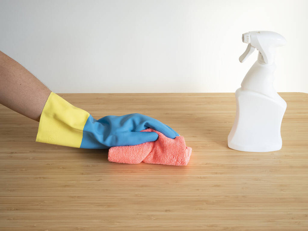 Manos caucásicas limpiando una mesa de madera con guantes azules y un spray desinfectante blanco en cualquier tarde dada. - Foto, imagen