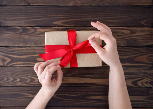 Verbeugung im Geschenkkarton. Frau wickelt Urlaub handgemachtes Geschenk in Papier mit roter Schleife auf Holztisch, Ansicht von oben - Foto, Bild