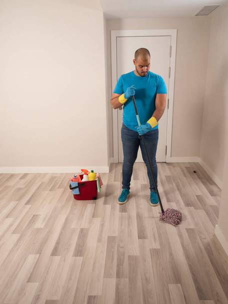 Joven hombre limpieza con un cepillo de barrido y fregona y cubo de agua y artículos de limpieza en casa con guantes azules y camisa azul - Foto, Imagen