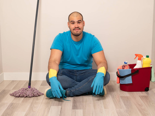 Junger Mann putzt mit Kehrbürste und Wischmopp und Eimer Wasser und Putzmittel zu Hause mit blauen Handschuhen und blauem Hemd - Foto, Bild