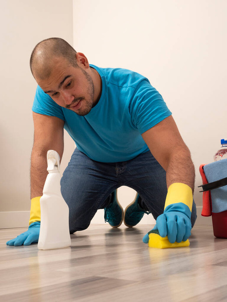 Νεαρός καθαρίζει με βούρτσα και σφουγγαρίστρα και κουβά νερό και προμήθειες καθαρισμού στο σπίτι φορώντας μπλε γάντια και μπλε πουκάμισο - Φωτογραφία, εικόνα