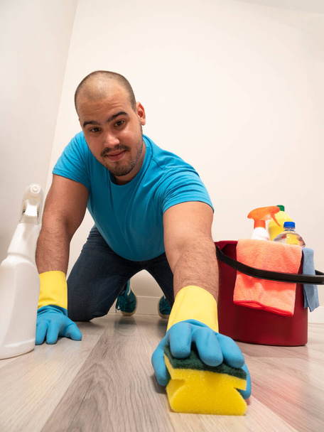 Young man cleaning with a sweeping brush and mop and bucket of water and cleaning supplies at home wearing blue gloves and blue shirt - Zdjęcie, obraz
