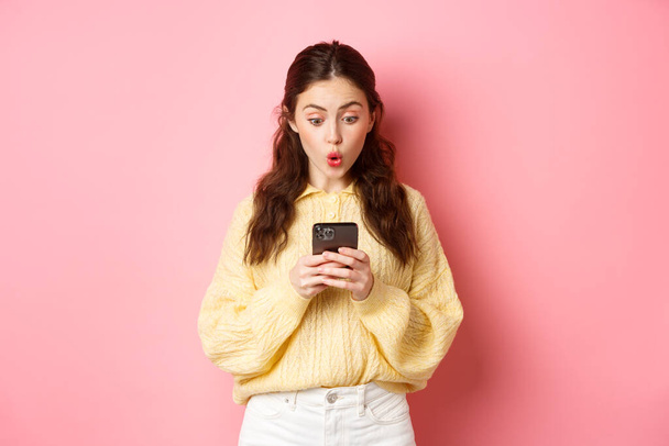 Portrait of surprised young woman reading message on smartphone and say wow, look curious at mobile screen, standing against pink background - Фото, изображение