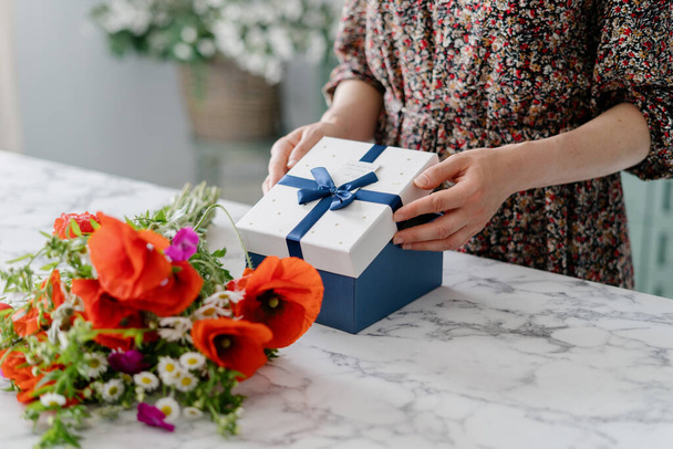 Cropped shot of woman wearing dress opening gift box with blue ribbon and bow, taking white top off, peeking inside, marble table with colorful flower bouquet, blurred background - Photo, Image