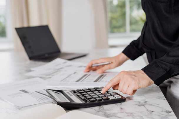 Πρόσωπο με μαύρο πουκάμισο που υπολογίζει τους μηνιαίους λογαριασμούς και τα χρέη με τη βοήθεια της αριθμομηχανής και laptop, τιμολόγια που βρίσκονται γύρω από το μαρμάρινο τραπέζι με παράθυρα σε θολή φόντο. Καλλιεργημένη βολή - Φωτογραφία, εικόνα