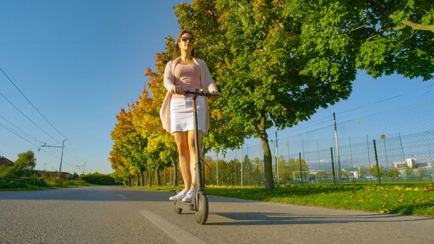 LOW ANGLE: Young woman commutes along a fall colored avenue on an e-scooter. - Foto, Bild