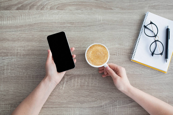 Modern lifestyle. Woman holsing smartphone with blank black screen and drinking coffee at workplace, top view - Photo, Image