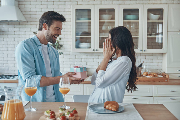 νεαρός άνδρας δίνει ένα κουτί δώρου στην κοπέλα του, ενώ παίρνει πρωινό στην οικιακή κουζίνα - Φωτογραφία, εικόνα