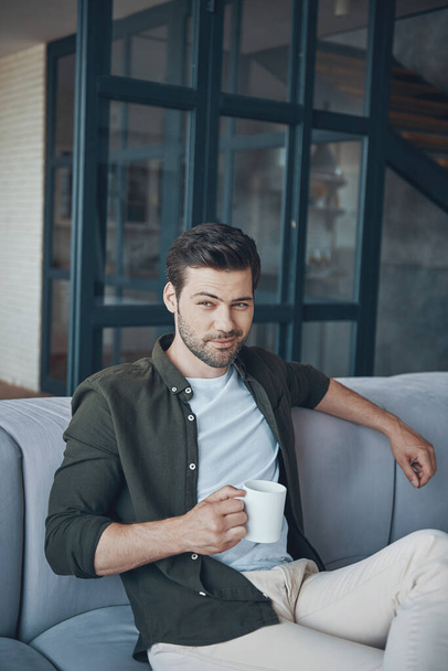Handsome young man holding cup with drink while sitting on the sofa at home - Photo, image