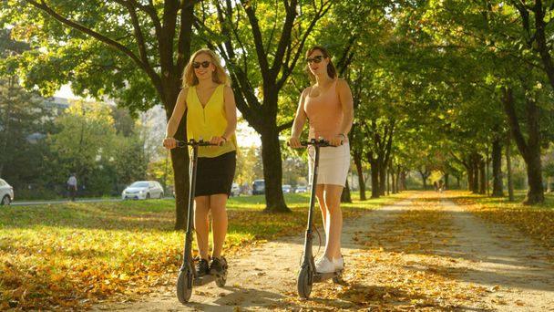 Dos amigas felices hablan mientras montan e-scooters alrededor del parque de color otoño - Foto, Imagen