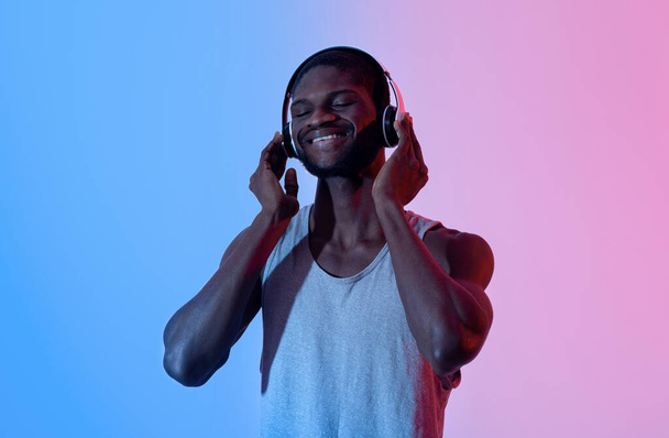 Черный спортсмен, используя современные наушники, слушая классные саундтреки, выбирая плейлист для тренировки в неоновом свете - Фото, изображение