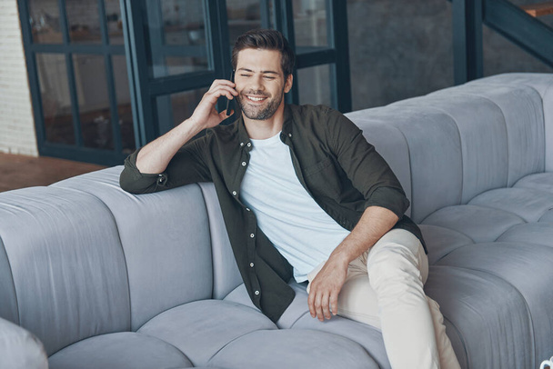 Handsome young man talking on smart phone and looking away while sitting on the sofa at home - Foto, Imagen