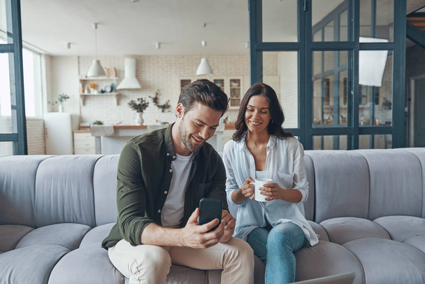 Hermosa pareja joven bebiendo café y comunicándose mientras se sienta en el sofá en casa - Foto, imagen