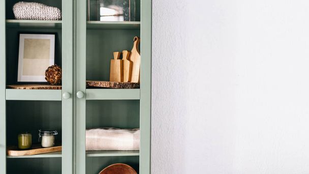 Tall green cabinet with glass doors and various kitchen equipment on shelves such as wooden cutting boards and tablecloths inside, vintage furniture, white wall in background. Copy space for text - 写真・画像