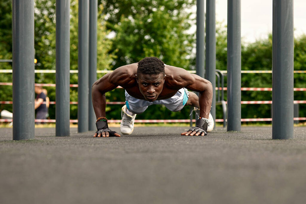 Urheilukentällä treenaava lihaksikas nuori mies. Afrikkalainen mies etsii sivuun tehdessään vaakasuora baari harjoituksia. Paidaton miesmalli käyttää ulkona. - Valokuva, kuva