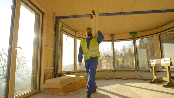 LOW ANGLE: Pracownik przychodzi do pracy niosąc ciężkie drewniane belki na ramieniu - Zdjęcie, obraz
