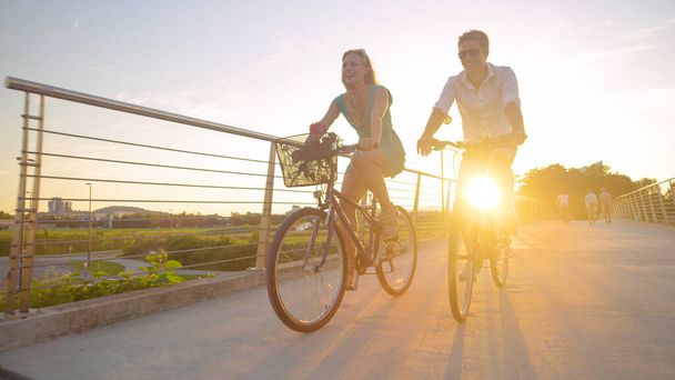 LENS FLARE Mujer joven sonríe mientras monta en bicicleta con su novio al amanecer - Foto, Imagen