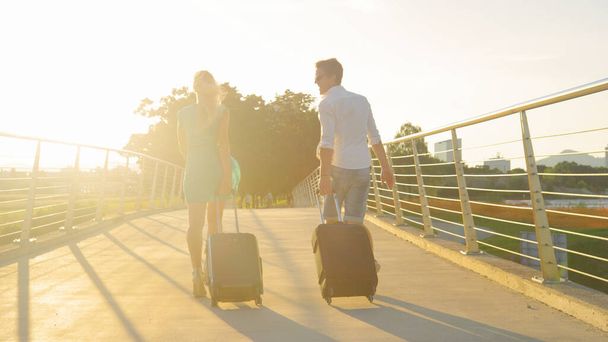 SUN FLARE: Mladý turistický pár kráčí přes nadjezd se svými zavazadly. - Fotografie, Obrázek