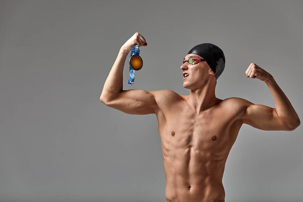Пловец с медалью на сером фоне радуется победе, спортсмен в отличной физической форме празднует победу, сжимая медаль в руке, победную концепцию, серый фон, пространство для копирования - Фото, изображение
