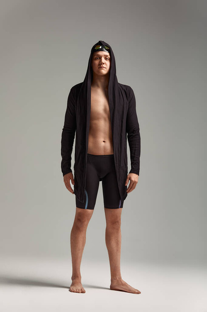 Guy nadador em um boné, máscara e roupão de banho para nadar, em um fundo cinza, se preparando para o nado - Foto, Imagem