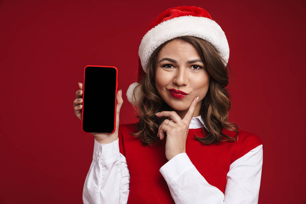 Image of thinking displeased young woman in christmas santa hat showing display of mobile phone isolated over red wall background - Zdjęcie, obraz