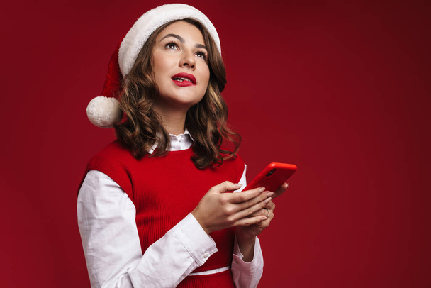 Photo of thinking young woman in christmas santa hat using mobile phone isolated over red wall background - Fotó, kép