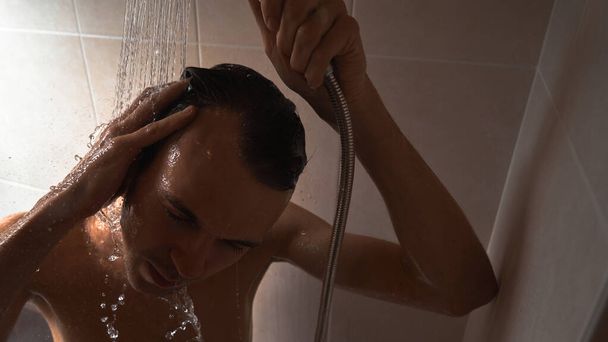 Portrait of young handsome man washes himself with shower gel, lathers head with shampoo in the bathroom at home close-up - Фото, зображення