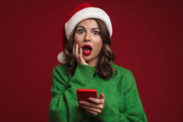 Photo of shocked young woman in christmas santa hat using mobile phone isolated over red wall background - Фото, изображение