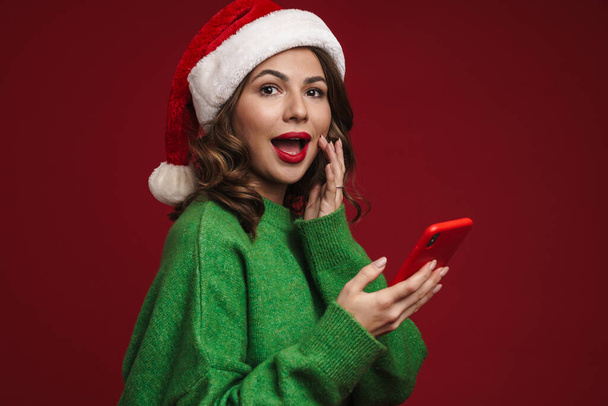 Изображение счастливой молодой женщины в рождественской шляпе Санты с помощью мобильного телефона на фоне красной стены - Фото, изображение