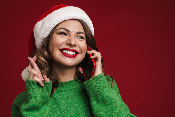 Photo of cheerful young woman in christmas santa hat talking by mobile phone and showing crossed fingers gesture isolated over red wall background - Foto, Imagem