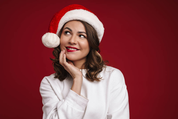 Photo of positive dreaming young woman in christmas santa hat isolated over red wall background - Foto, Imagem