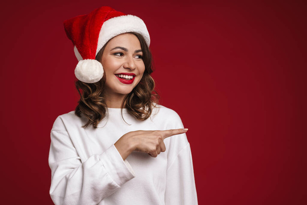 Фото щасливої веселої молодої жінки в різдвяному капелюсі Санта ізольовано на фоні червоної стіни, показуючи копійку
 - Фото, зображення