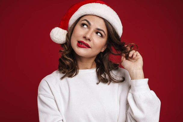 Image of dreaming young woman in christmas santa hat isolated over red wall background - Φωτογραφία, εικόνα