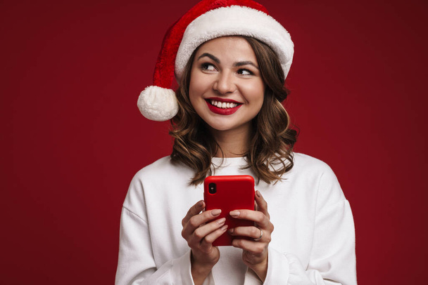 Bild von fröhlich glücklich junge Frau in Weihnachtsmann Hut mit Handy isoliert über rote Wand Hintergrund - Foto, Bild