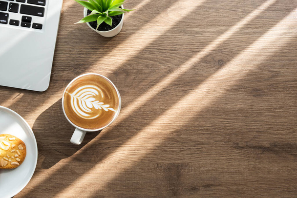 Ξύλινο τραπέζι γραφείου με φορητό υπολογιστή, ένα πιάτο μπισκότα αμυγδάλου και ένα φλιτζάνι καφέ latte με πρωινό ηλιακό φως. Πρωινό σνακ έννοια. Κάτοψη με χώρο αντιγραφής, επίπεδη lay. - Φωτογραφία, εικόνα