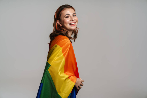 Happy beautiful woman smiling while posing with rainbow flag isolated over gray background - Photo, Image