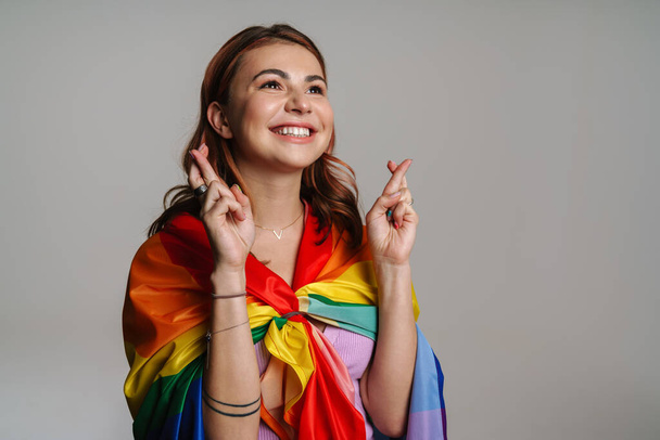 Cheerful woman wearing rainbow flag holding fingers crossed for good luck isolated over gray background - Foto, afbeelding