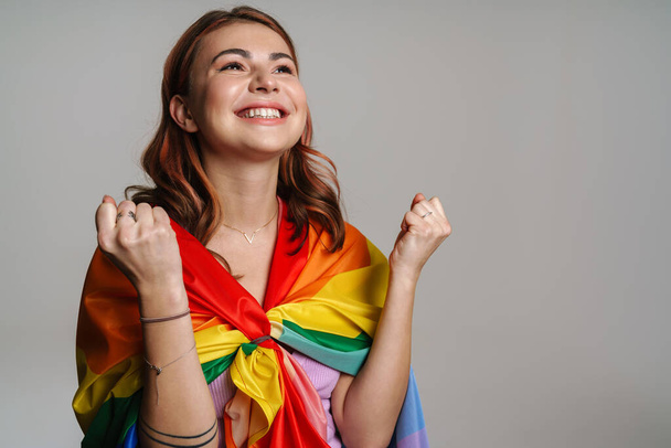 Joyful woman making winner gesture while posing with rainbow flag isolated over gray background - Foto, Imagem
