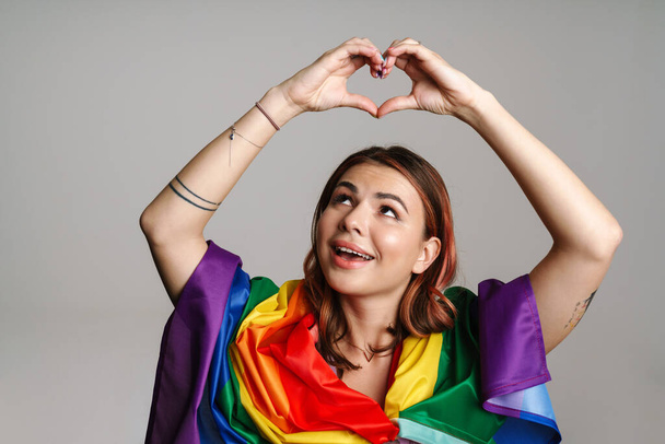 Cheerful woman wearing rainbow flag smiling and showing heart gesture isolated over gray background - Foto, Imagem