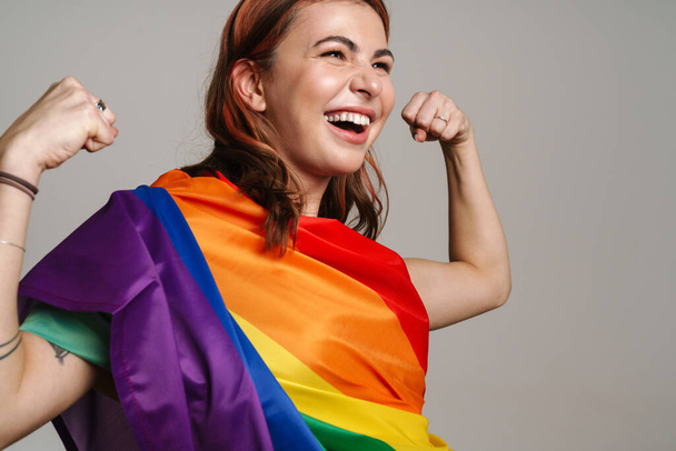 Cheerful woman wearing rainbow flag laughing and showing her biceps isolated over gray background - Фото, зображення