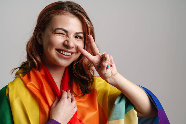 Cheerful woman wearing rainbow flag winking and showing peace sign isolated over gray background - Foto, afbeelding