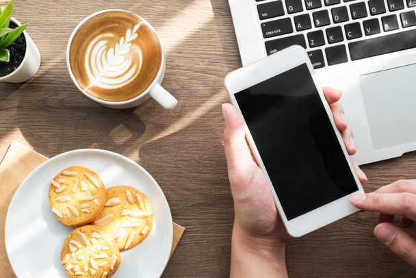 Smartphone de mano con pantalla de simulacro en blanco sobre mesa de escritorio de oficina de madera con galletas, taza de café y suministros. Vista superior, plano. - Foto, imagen